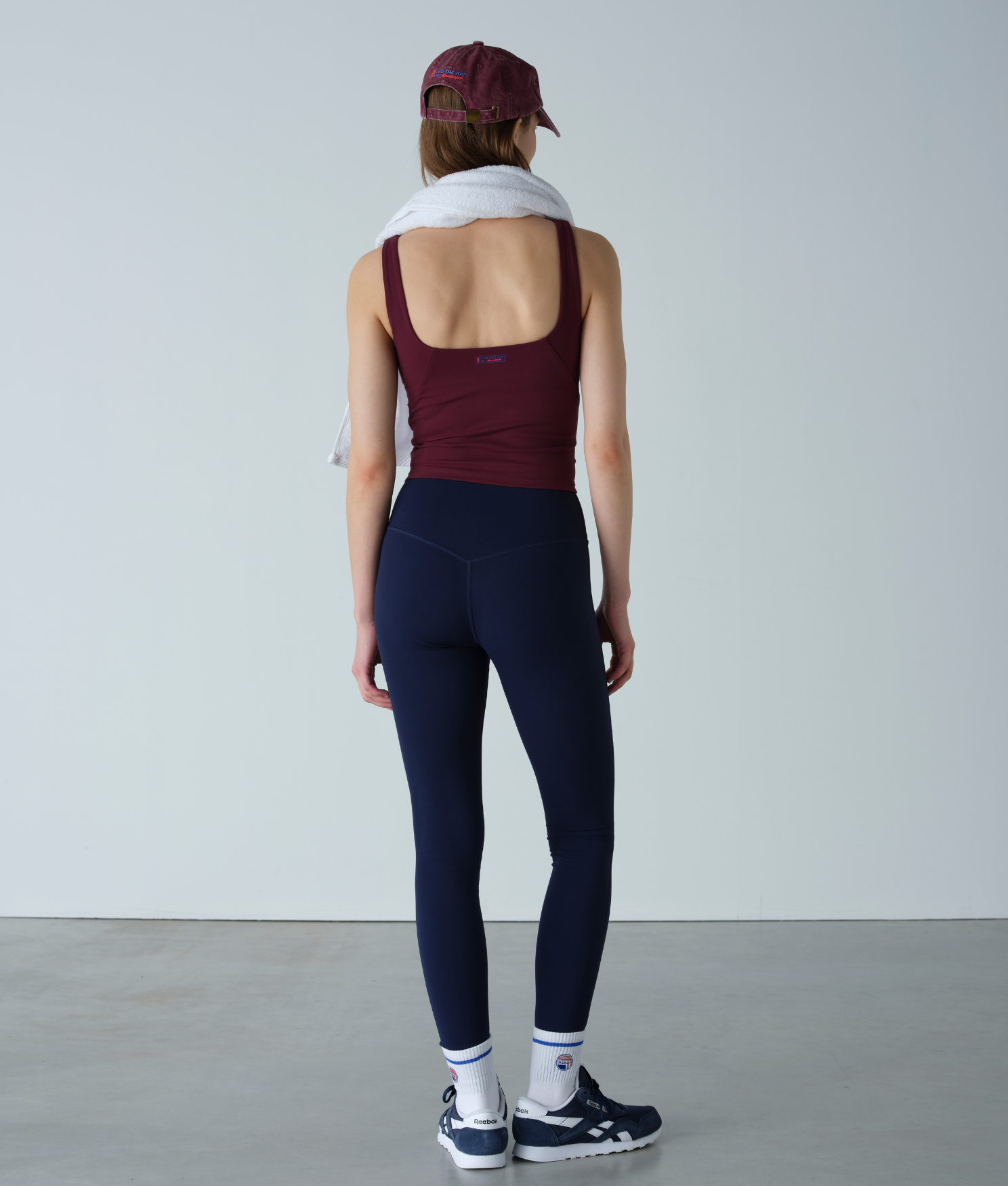 Person wearing a maroon tank top and navy leggings with a white towel over their shoulder against a plain background.